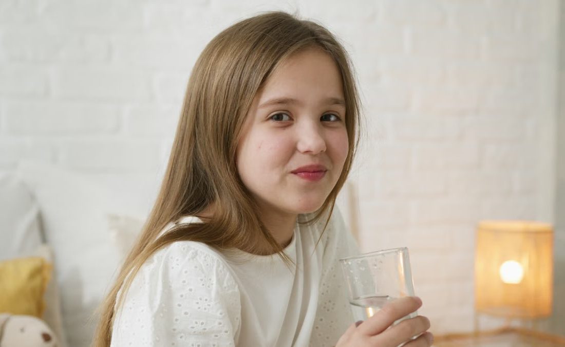 A young girl smiling and holding a glass of water inside a cozy bedroom setting.