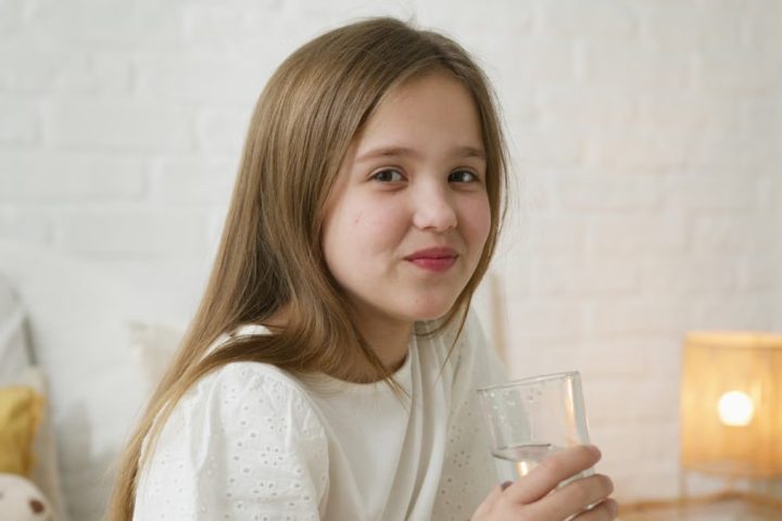 A young girl smiling and holding a glass of water inside a cozy bedroom setting.