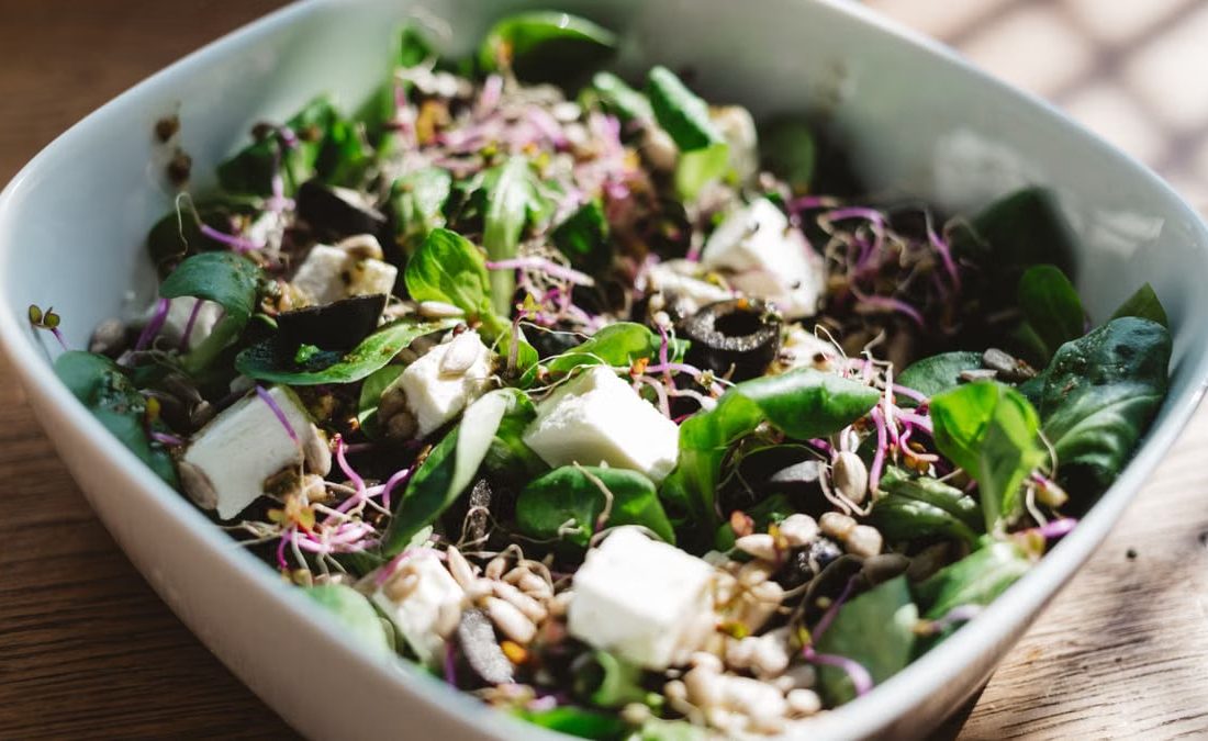 Fresh spinach salad with feta, olives, and sprouts, served in a sunlit bowl.