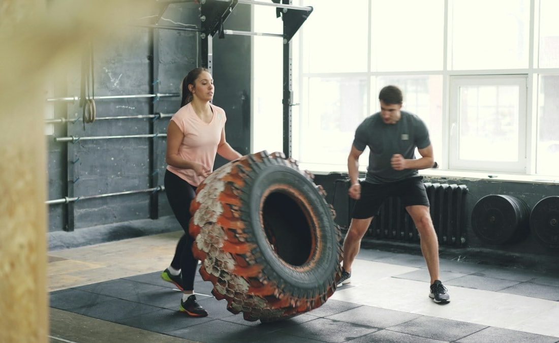 Girl in sports outfit is lifting and flipping heavy rubber tyre working out with coach in gym enjoying sports training together. People and activity concept.