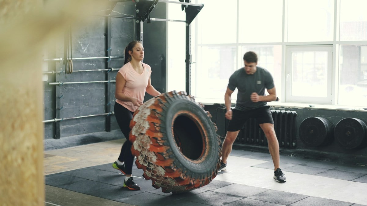 Girl in sports outfit is lifting and flipping heavy rubber tyre working out with coach in gym enjoying sports training together. People and activity concept.